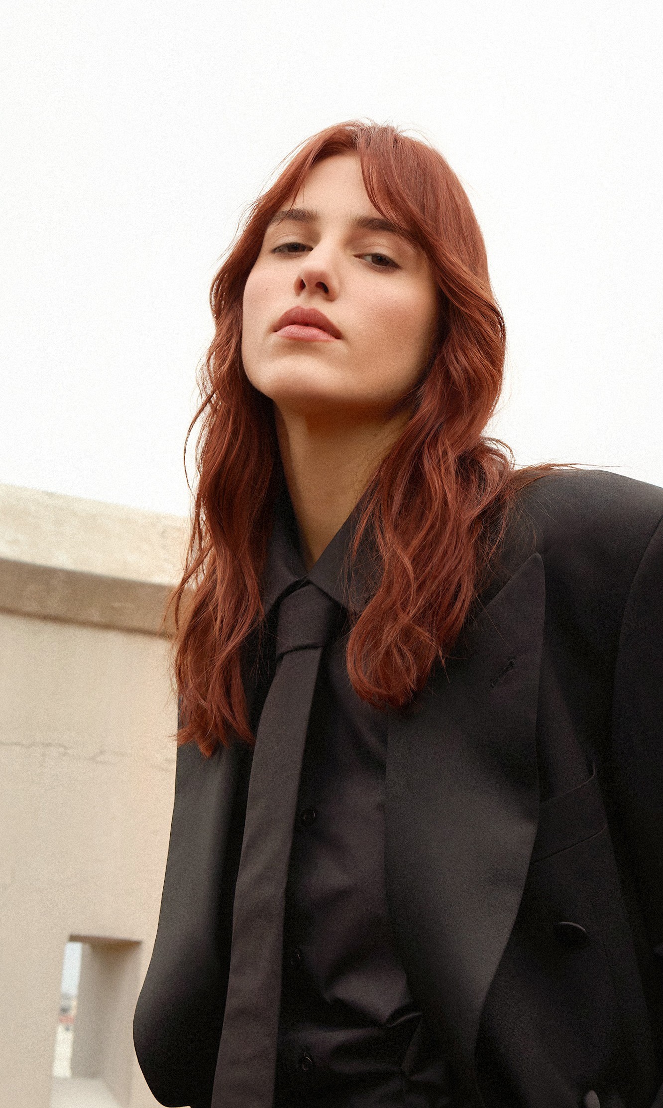 Musa Auburn Red: cabello largo cobrizo rojizo con flequillo, blazer y corbata negros, sobre una terraza de Buenos Aires.