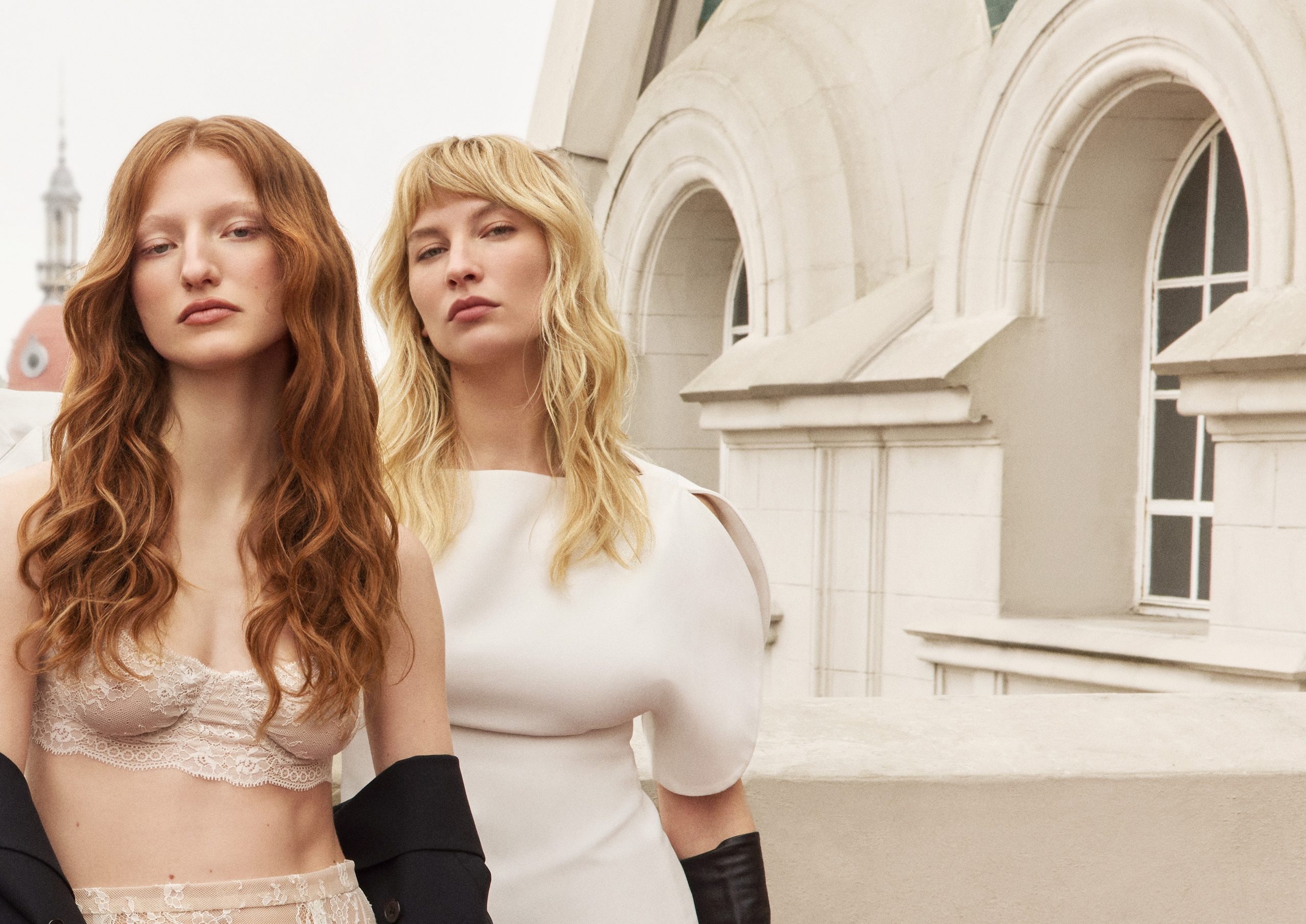 Dos musas, una de cabello cobrizo ondulado y otra rubia, junto a las cúpulas de un edificio histórico.