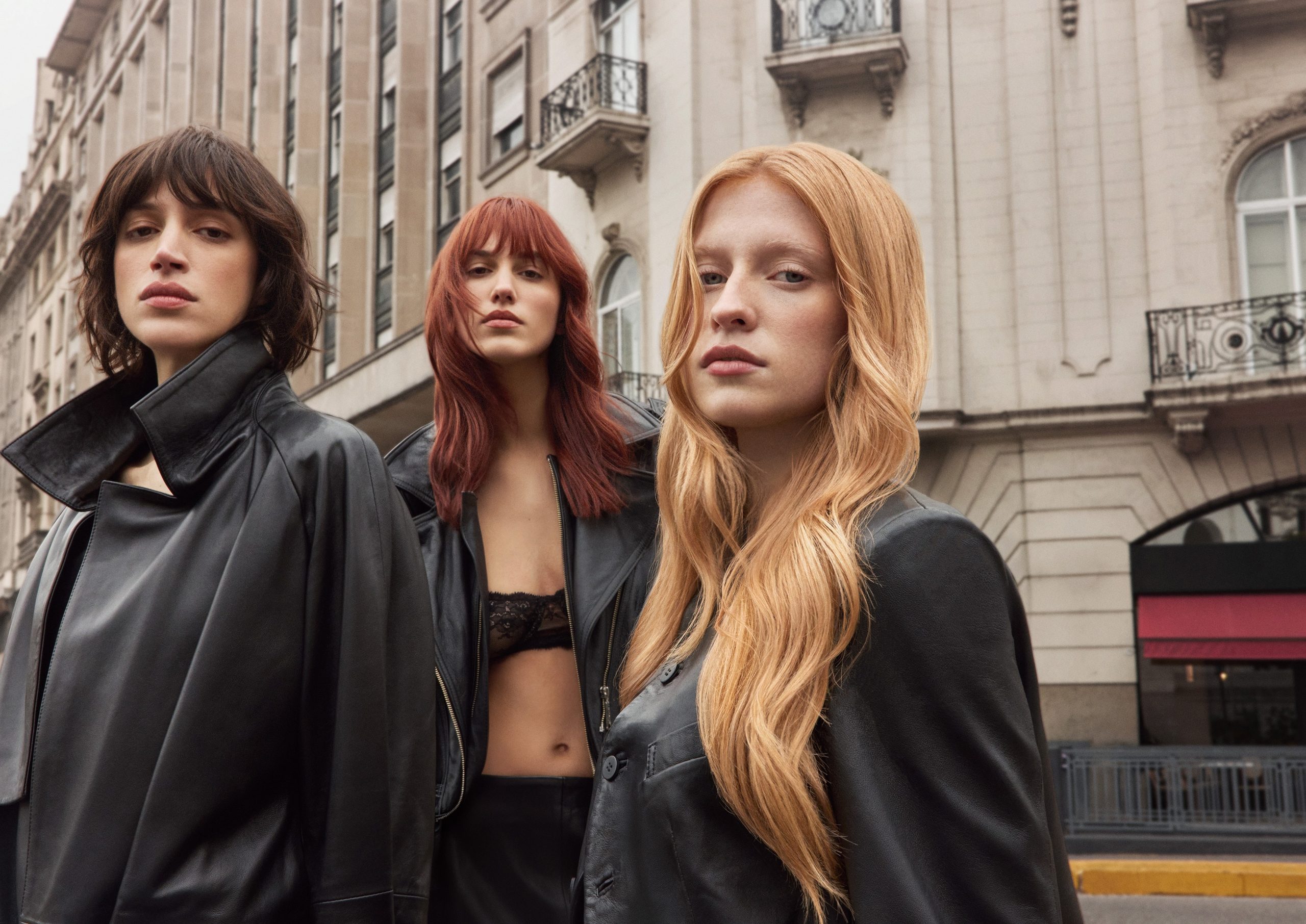 Tres musas con abrigos de cuero negro frente a una fachada clásica: castaño, cobrizo rojizo y rubio cobrizo.