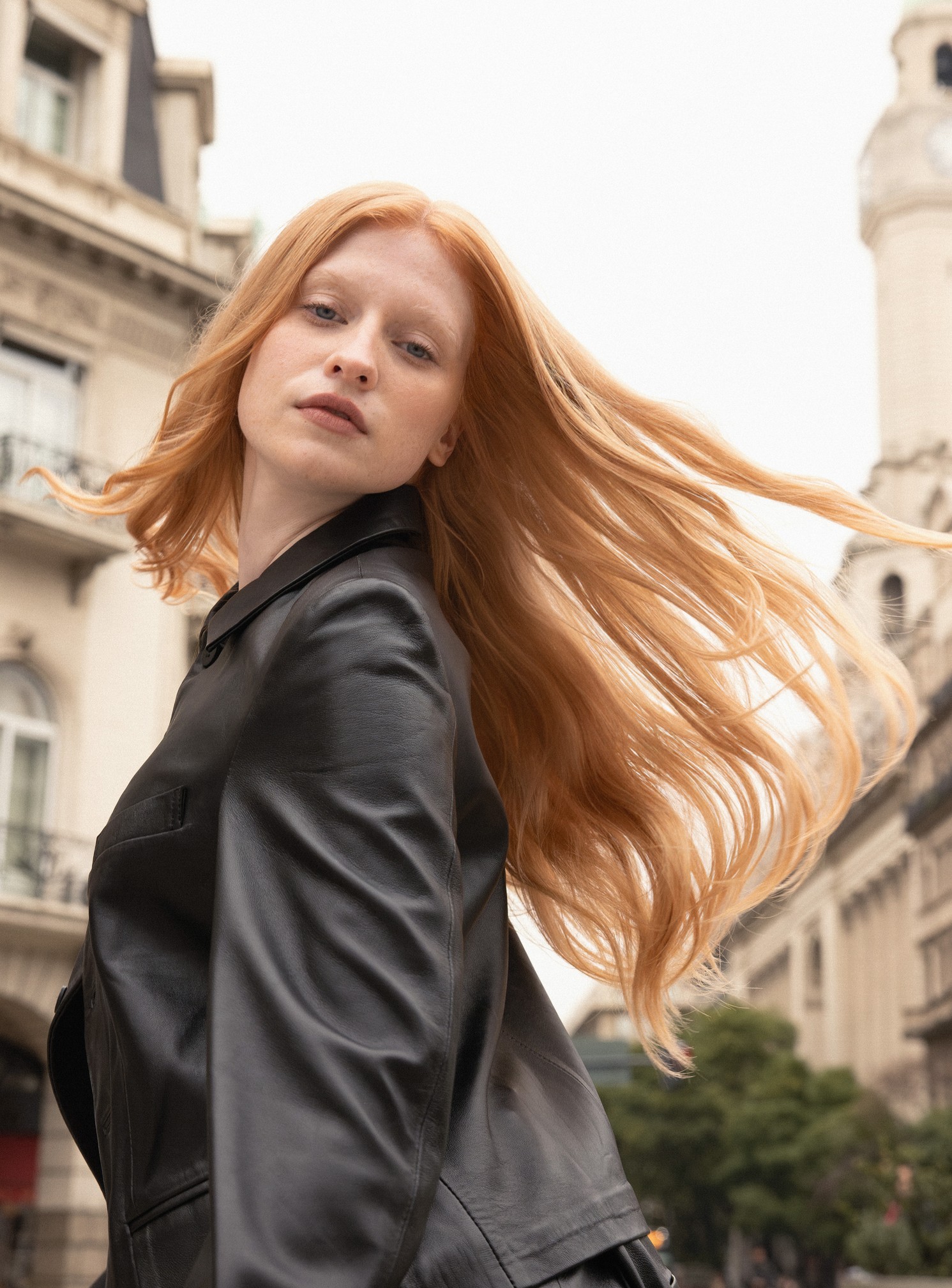 Musa de cabello largo rubio cobrizo movido por el viento, con campera de cuero, frente a edificios históricos de Buenos Aires.